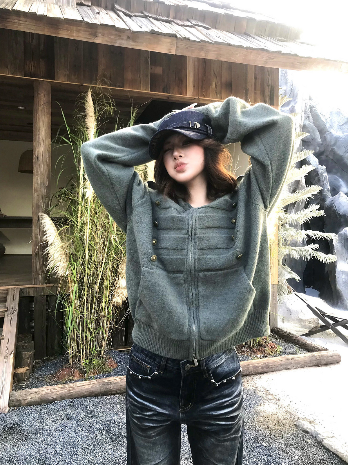 オリーブのニットジップパーカーを着た女性モデル。両手を頭に添えた上半身で、胸のボタンとショルダータブが見える
