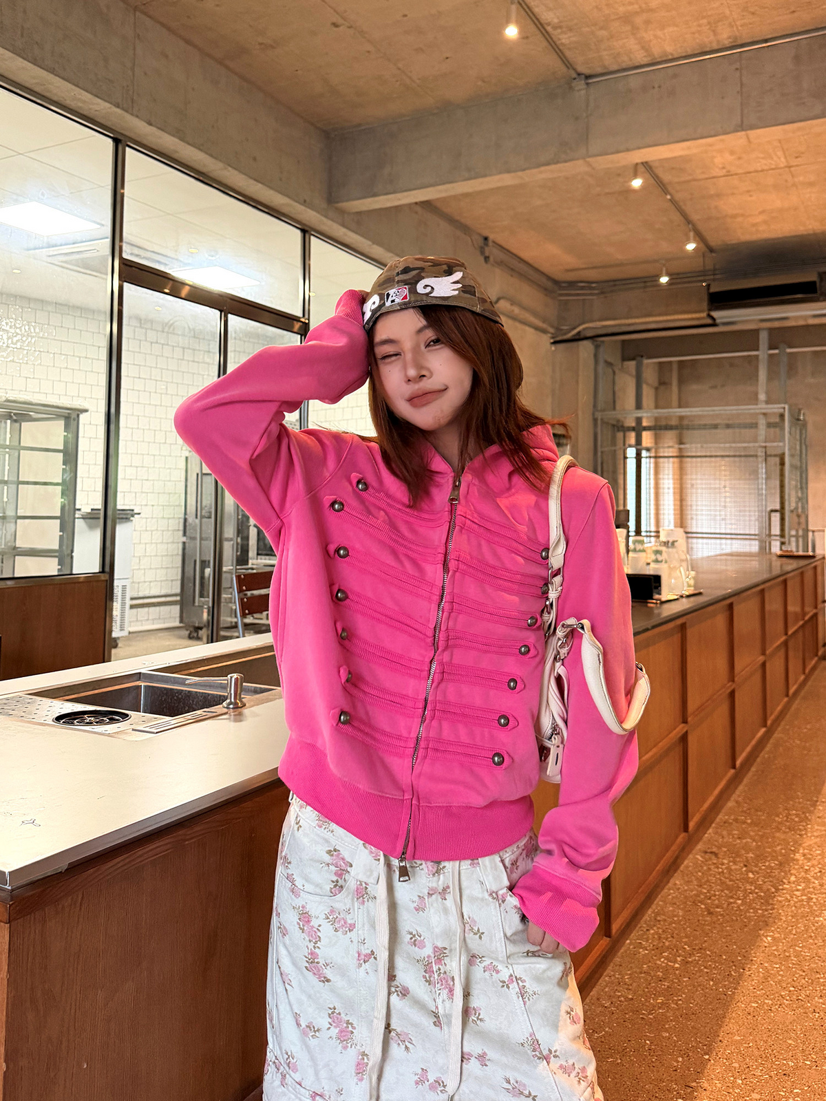 ホットピンクのミリタリー調ジップパーカーを着た女性モデル。片手を頭に添え、胸の飾り紐と金属ボタンが見える